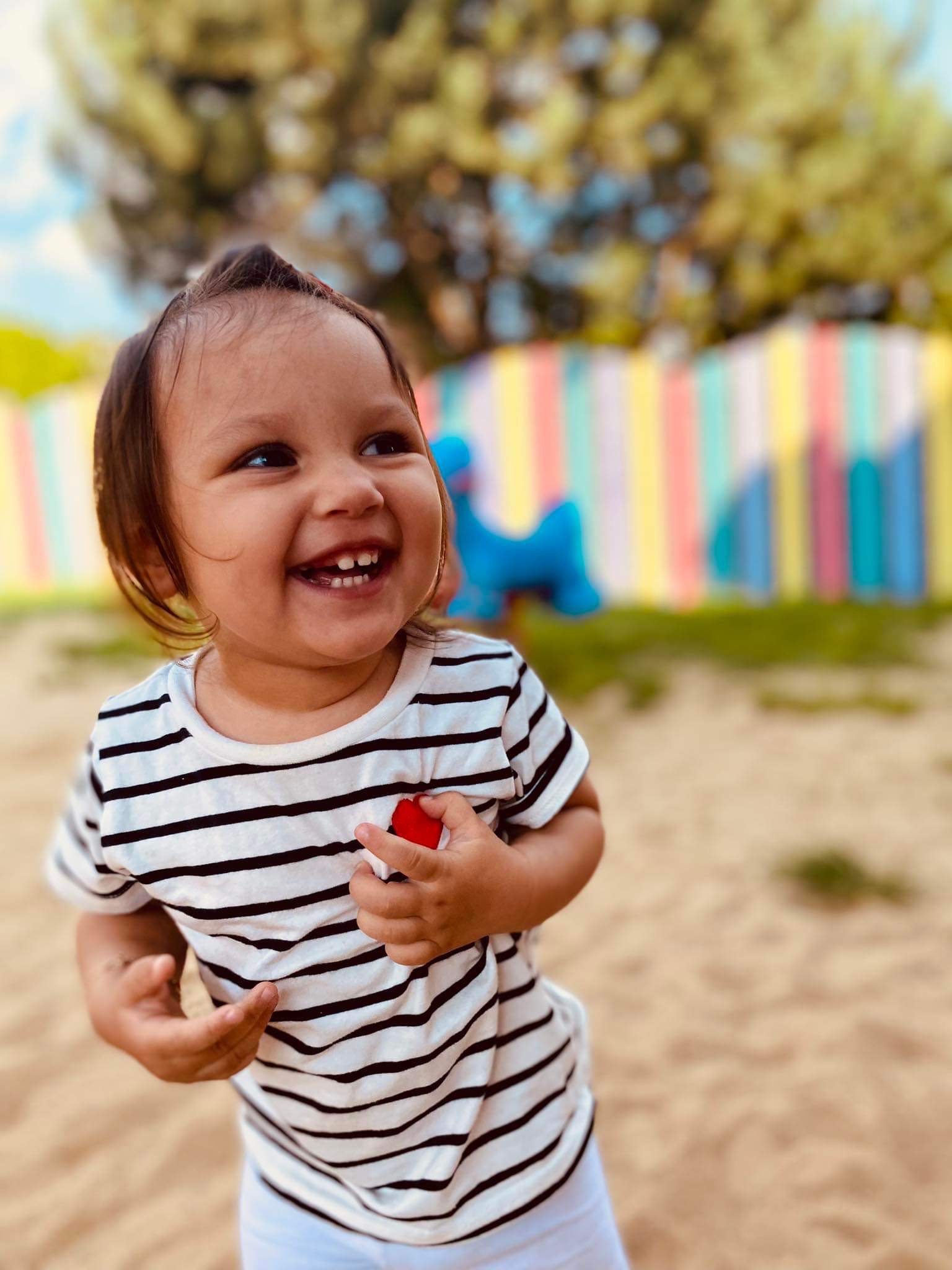Uśmiechnięta dziewczynka w pasiastym t-shircie, bawiąca się na piaszczystym placu zabaw z kolorowym płotem w tle, trzymająca w ręku czerwoną zabawkę.