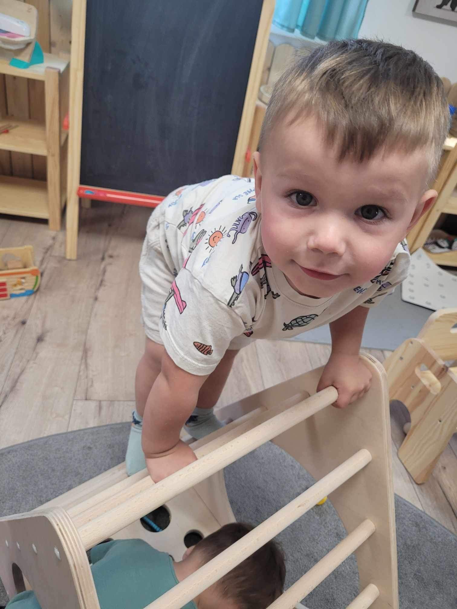 Chłopiec w koszulce z nadrukiem wspina się na drewniany trójkątny drabinkę w jasnym pomieszczeniu zabaw, z drugim dzieckiem widocznym pod konstrukcją.