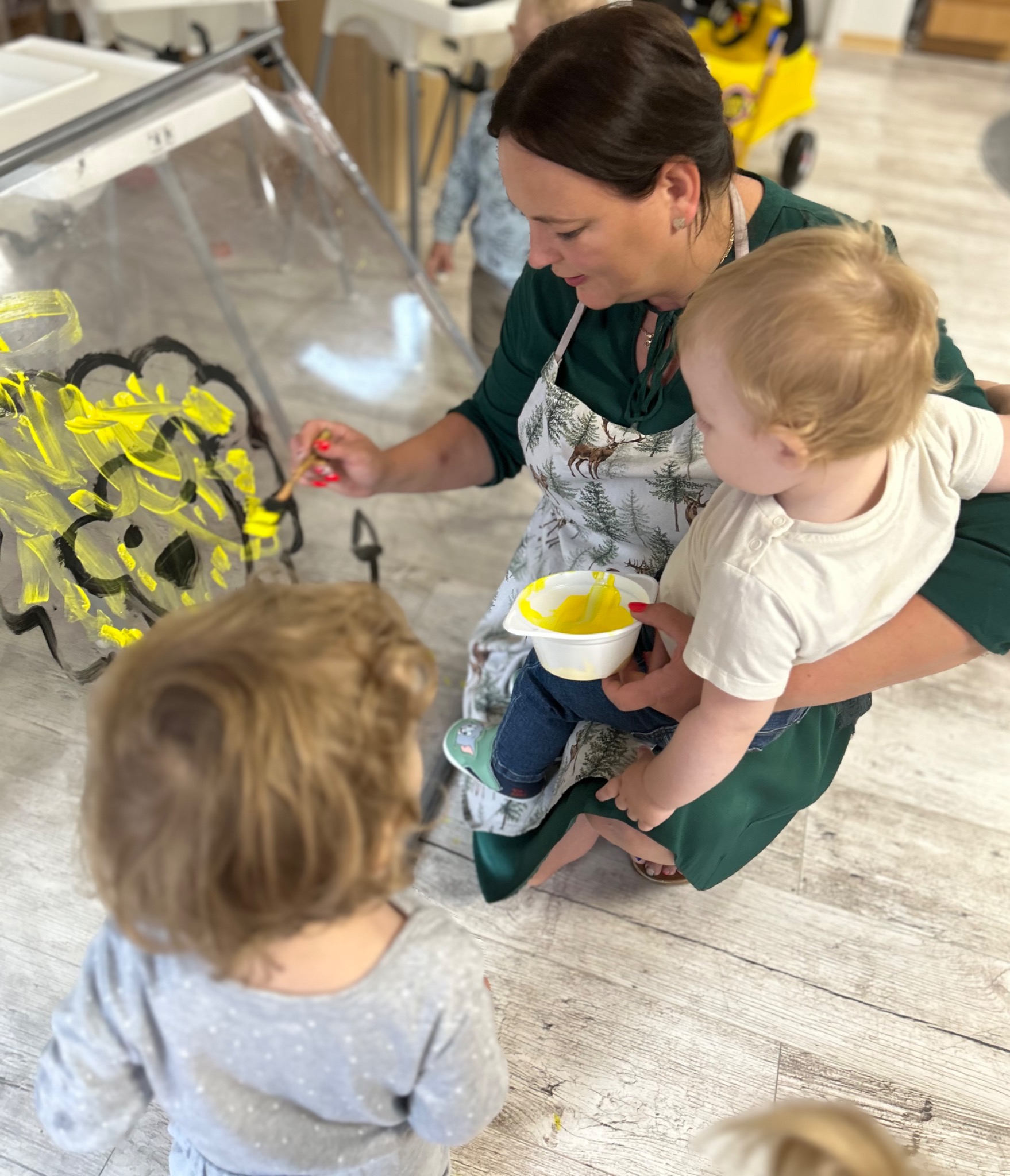 Kobieta w zielonej sukience i fartuchu z motywem leśnym maluje z dziećmi żółtą farbą na przezroczystej tablicy. Jedno dziecko trzyma na rękach, a drugie obserwuje z przodu.
