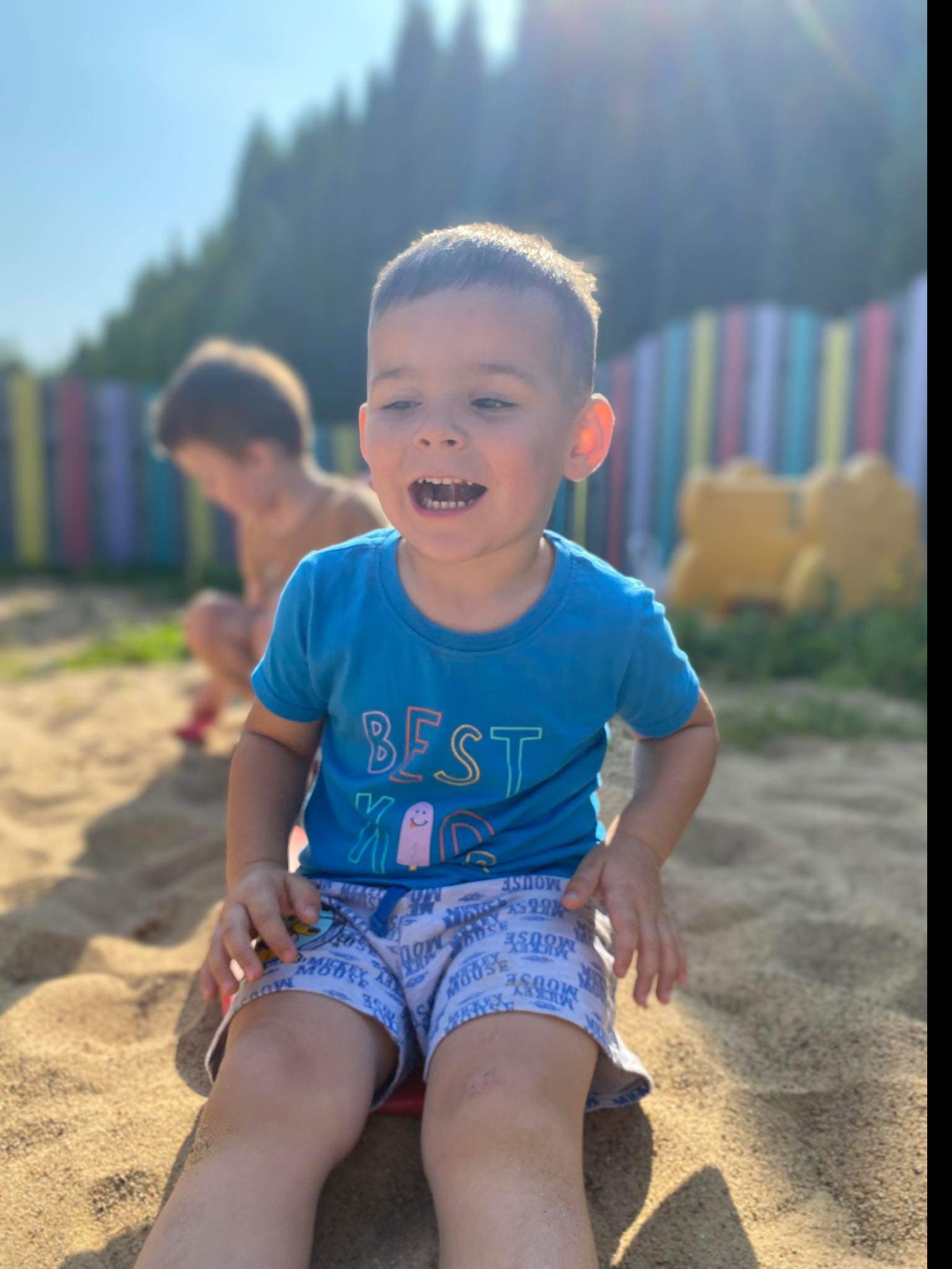 Uśmiechnięty chłopiec w niebieskiej koszulce z napisem 'BEST KID' siedzi w piaskownicy na placu zabaw w słoneczny dzień, w tle kolorowy płot i inne dziecko.