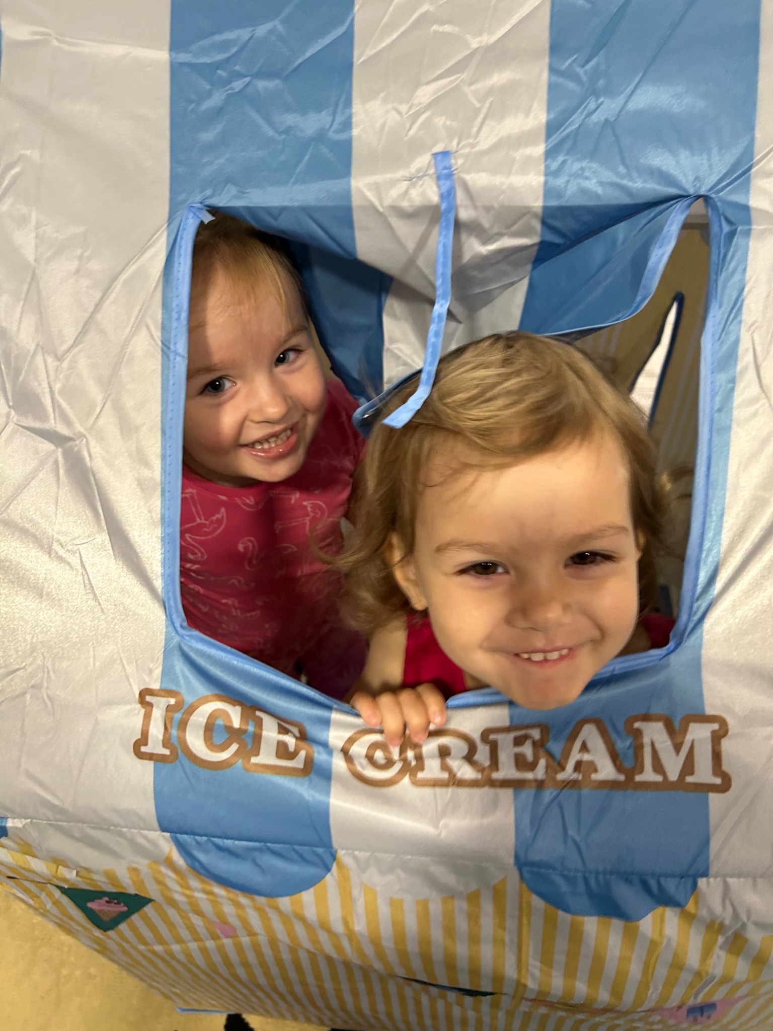 Dwie uśmiechnięte dziewczynki wyglądają z okna zabawkowego namiotu w kształcie budki z lodami, z widocznym napisem 'ICE CREAM'.