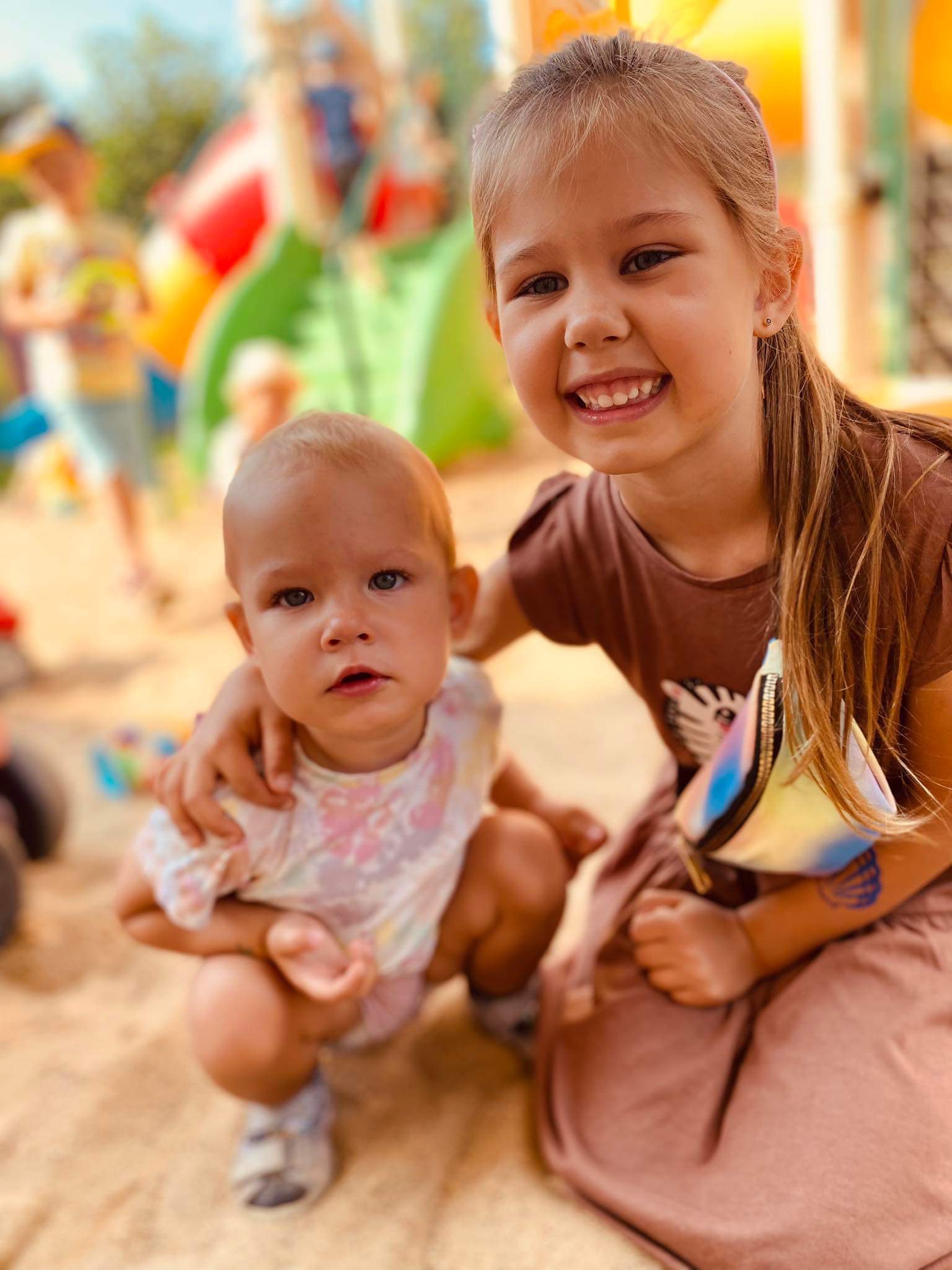 Uśmiechnięta dziewczynka obejmuje młodszą siostrę na tle kolorowego placu zabaw, obie siedzą w piasku.