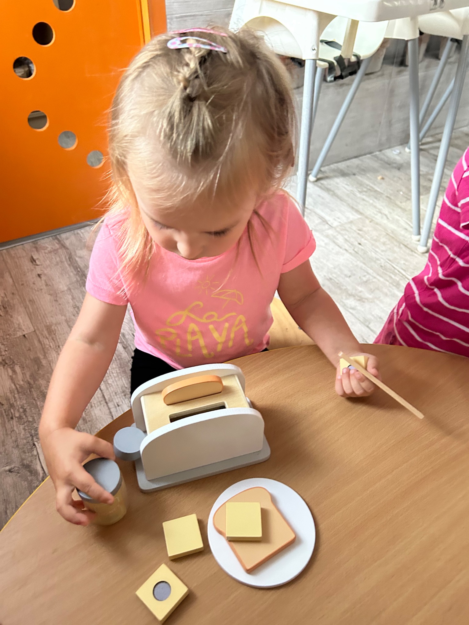 Dziewczynka z blond warkoczem bawi się drewnianym tosterem i akcesoriami do przygotowywania tostów na drewnianym stole.