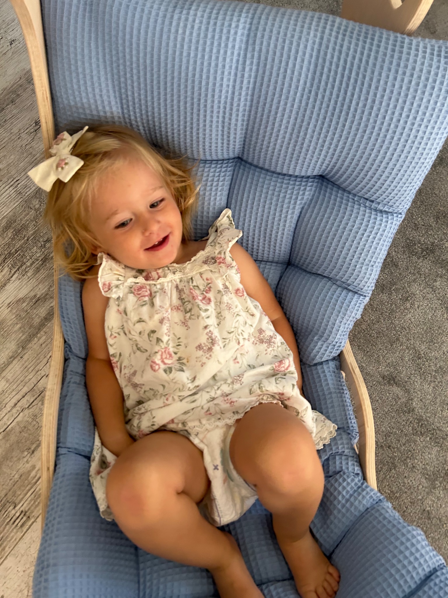 Uśmiechnięta blondwłosa dziewczynka w kwiecistej sukience i kokardą we włosach relaksuje się w błękitnym, miękkim fotelu bujanym.