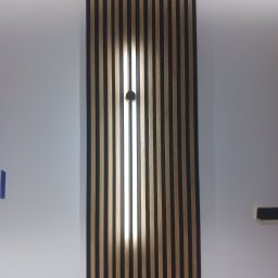 Elektro-max - Panel ścienny z drewnianych lameli, podświetlony od tyłu, z widocznym punktem mocowania oświetlenia w centralnej części.