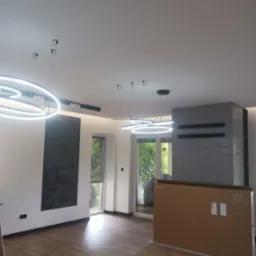 Nowoczesne oświetlenie LED w salonie z drewnianą podłogą, widoczne okrągłe lampy wiszące i karton z telewizorem Samsung OLED S85D 77 cali.