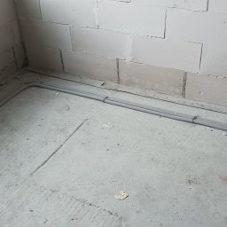 Elektro-max - Dwa szare karbowane przewody elektryczne ułożone wzdłuż kąta ściany z bloków betonowych na surowym betonowym podłożu w trakcie prac instalacyjnych.