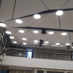 Elektro-max - Montaż sufitu podwieszanego z oświetleniem punktowym w dużej hali, widoczny pracownik na drabinie i rusztowania.
