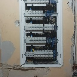 Elektro-max - Rozdzielnica elektryczna z licznymi zabezpieczeniami zamontowana w ścianie podczas remontu, widoczne przewody i fragmenty instalacji elektrycznej w surowym stanie.