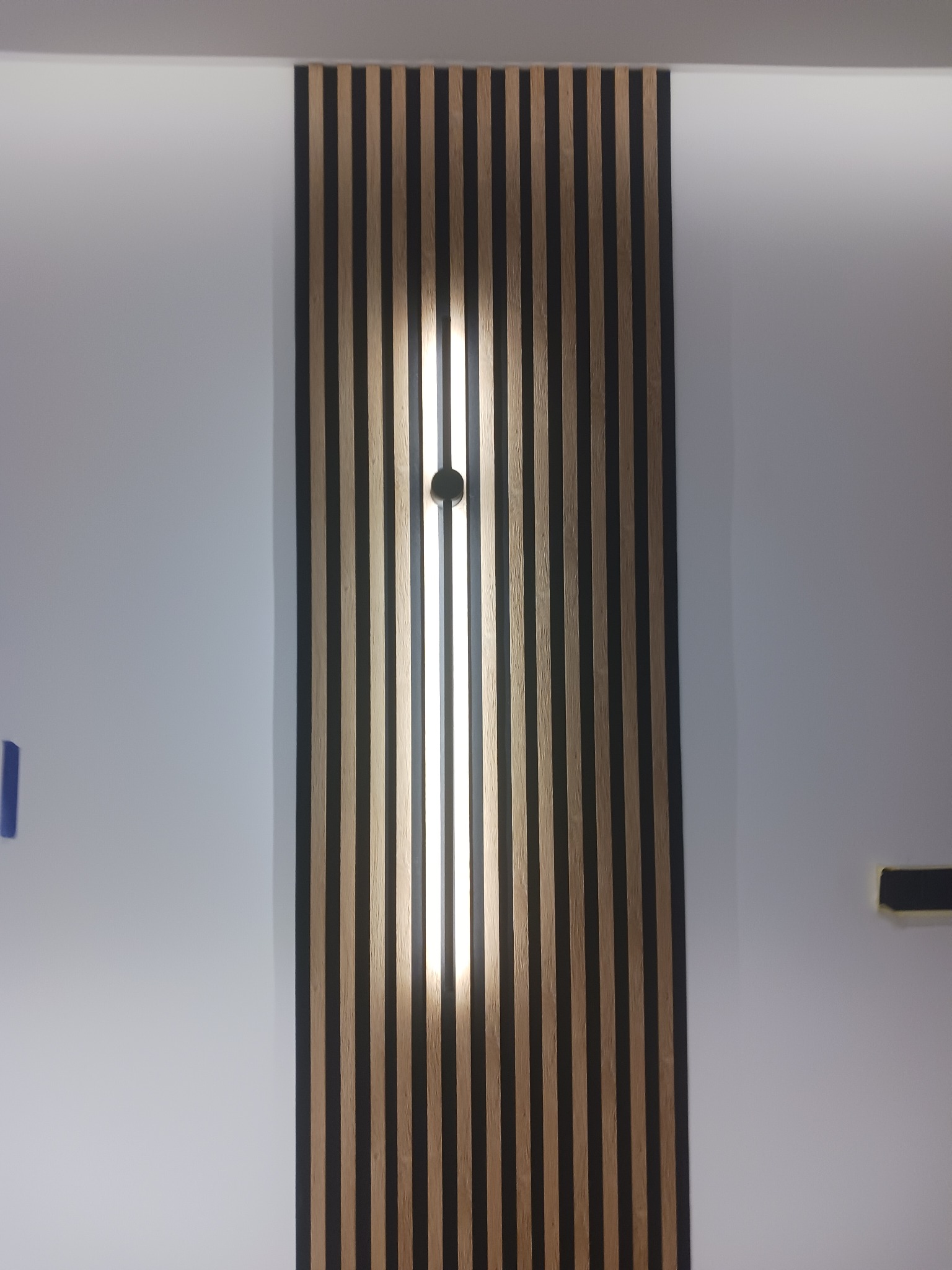 Panel ścienny z drewnianych lameli, podświetlony od tyłu, z widocznym punktem mocowania oświetlenia w centralnej części.