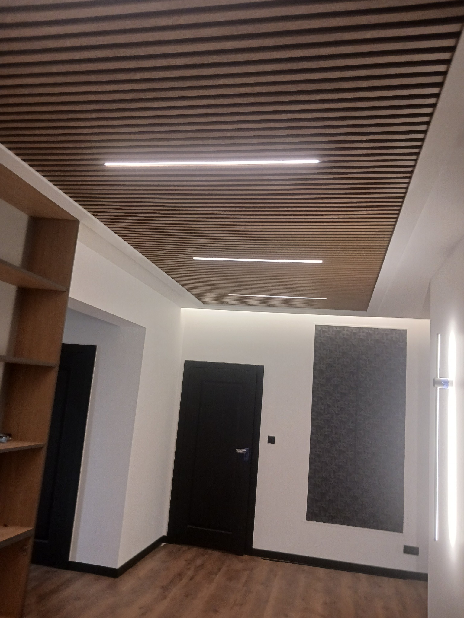 Nowoczesny korytarz z drewnianym sufitem lamelowym, oświetleniem LED wpuszczonym w sufit i pionowymi lampami ściennymi, uzupełniony ciemnymi drzwiami i wzorzystą tapetą.