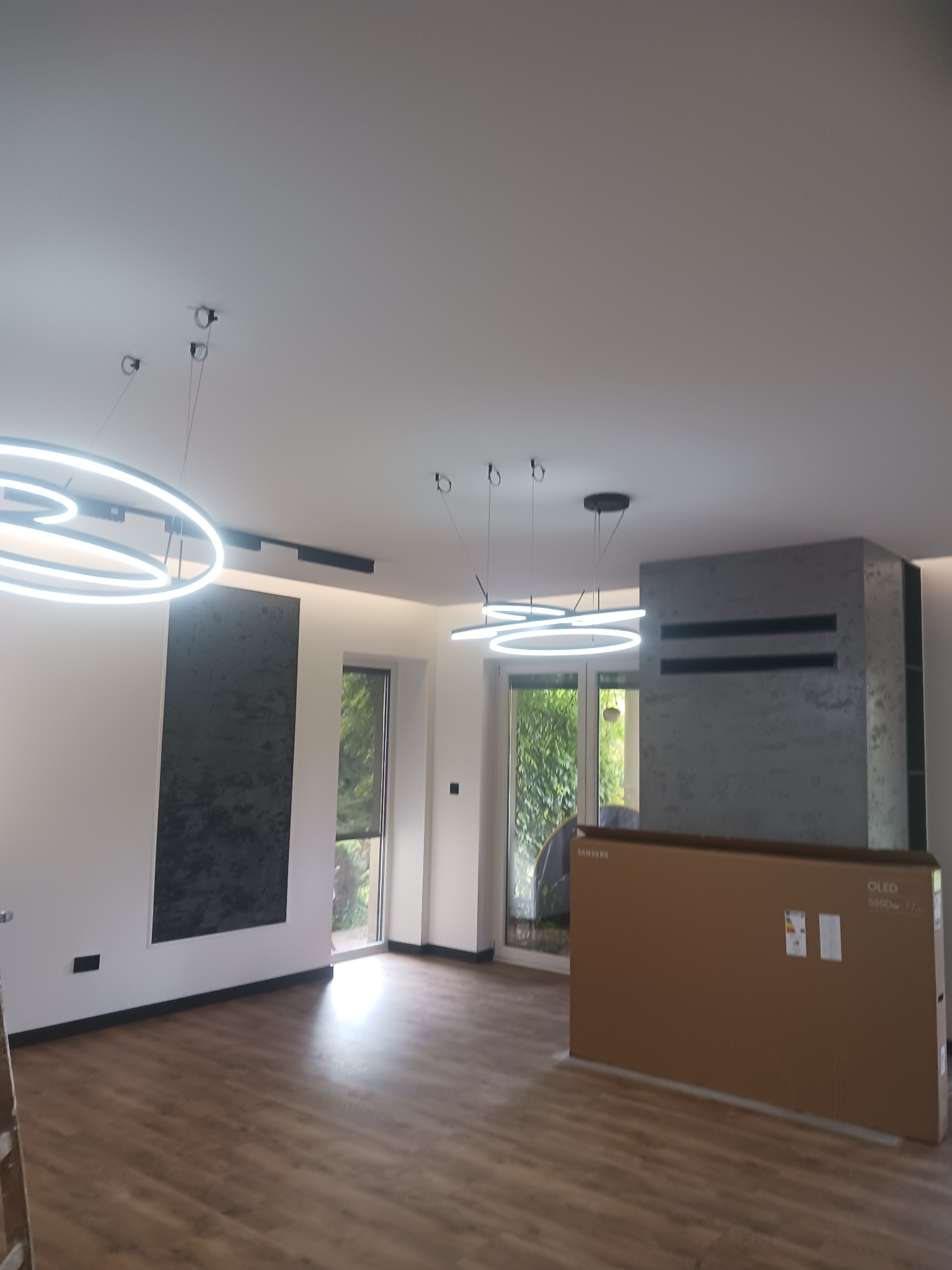 Nowoczesne oświetlenie LED w salonie z drewnianą podłogą, widoczne okrągłe lampy wiszące i karton z telewizorem Samsung OLED S85D 77 cali.