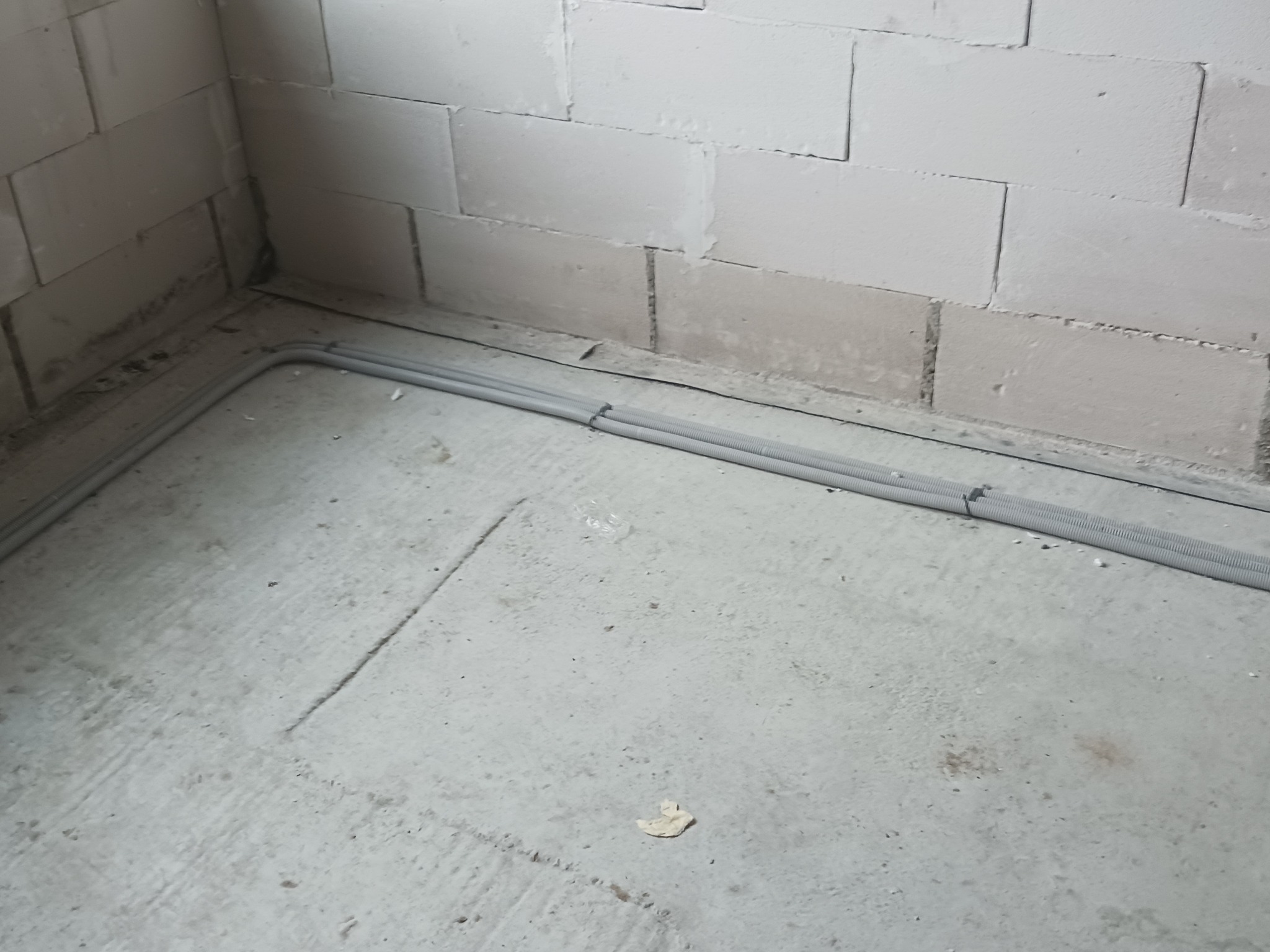 Dwa szare karbowane przewody elektryczne ułożone wzdłuż kąta ściany z bloków betonowych na surowym betonowym podłożu w trakcie prac instalacyjnych.