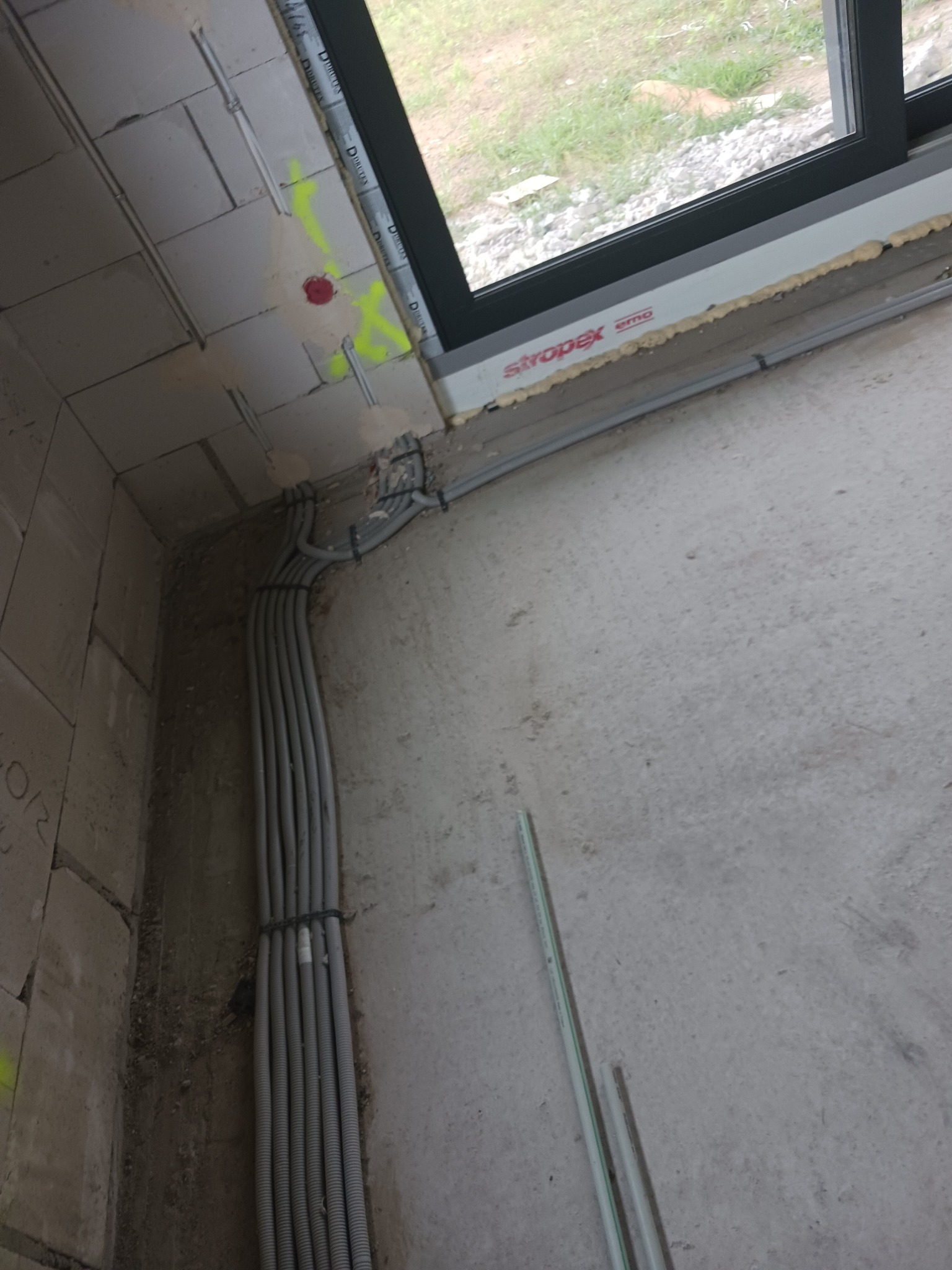 Wiązka szarych kabli elektrycznych prowadzona wzdłuż surowej ściany z betonu komórkowego, z widocznymi oznaczeniami w kolorze żółtym i czerwonym, w pobliżu okna z pianką montażową.