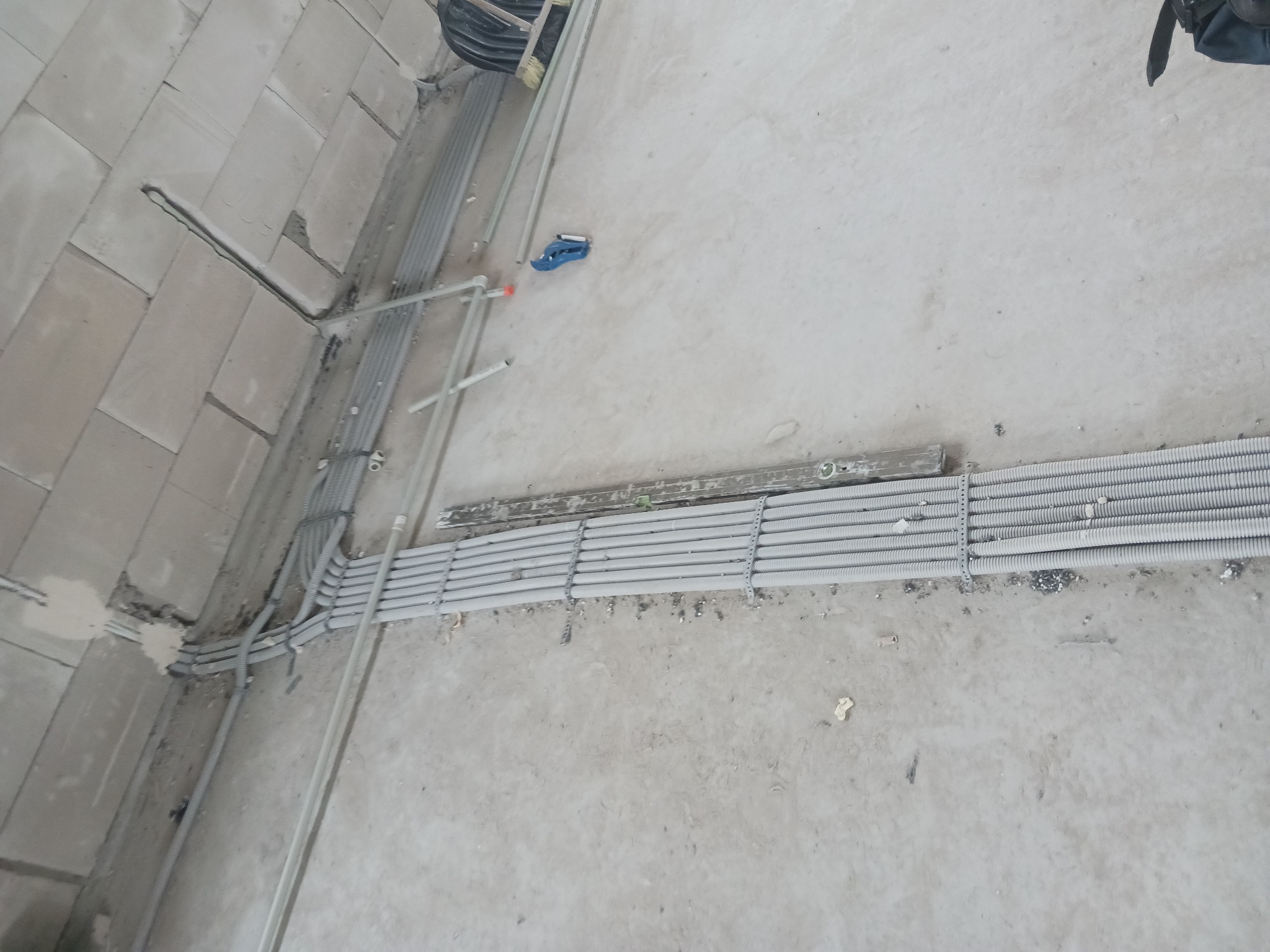 Szare rury karbowane ułożone równolegle na podłodze w surowym stanie, prowadzone wzdłuż ściany z betonu komórkowego, widoczne narzędzia i drabina.