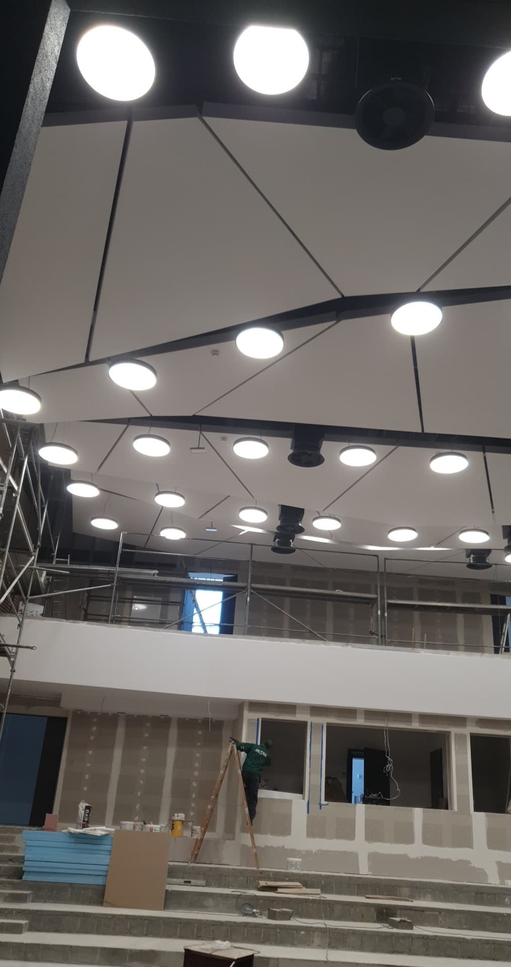 Montaż sufitu podwieszanego z oświetleniem punktowym w dużej hali, widoczny pracownik na drabinie i rusztowania.