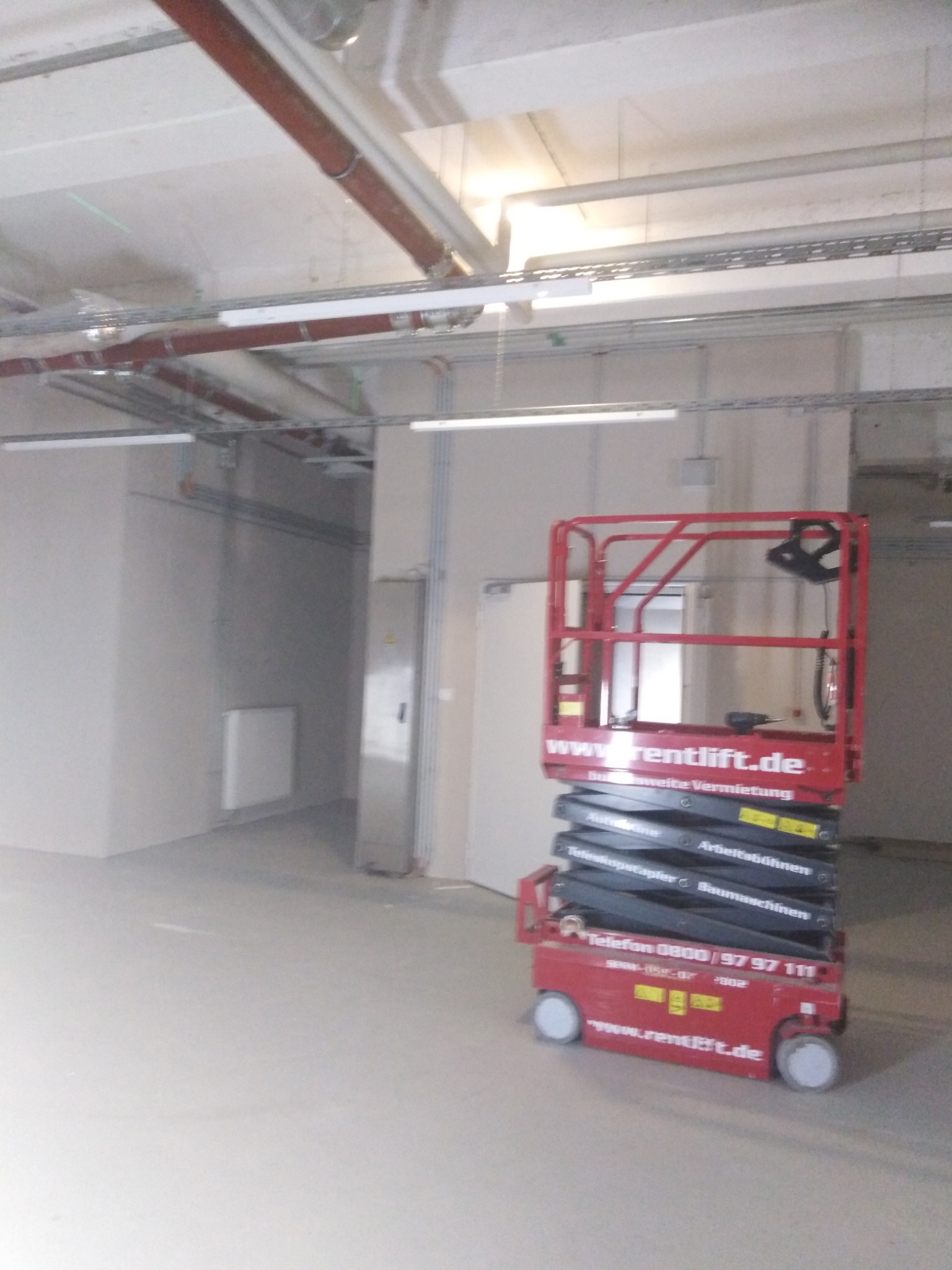 Wnętrze w trakcie prac instalacyjnych z widoczną podnośnikiem nożycowym, przewodami pod sufitem i skrzynką elektryczną na ścianie.