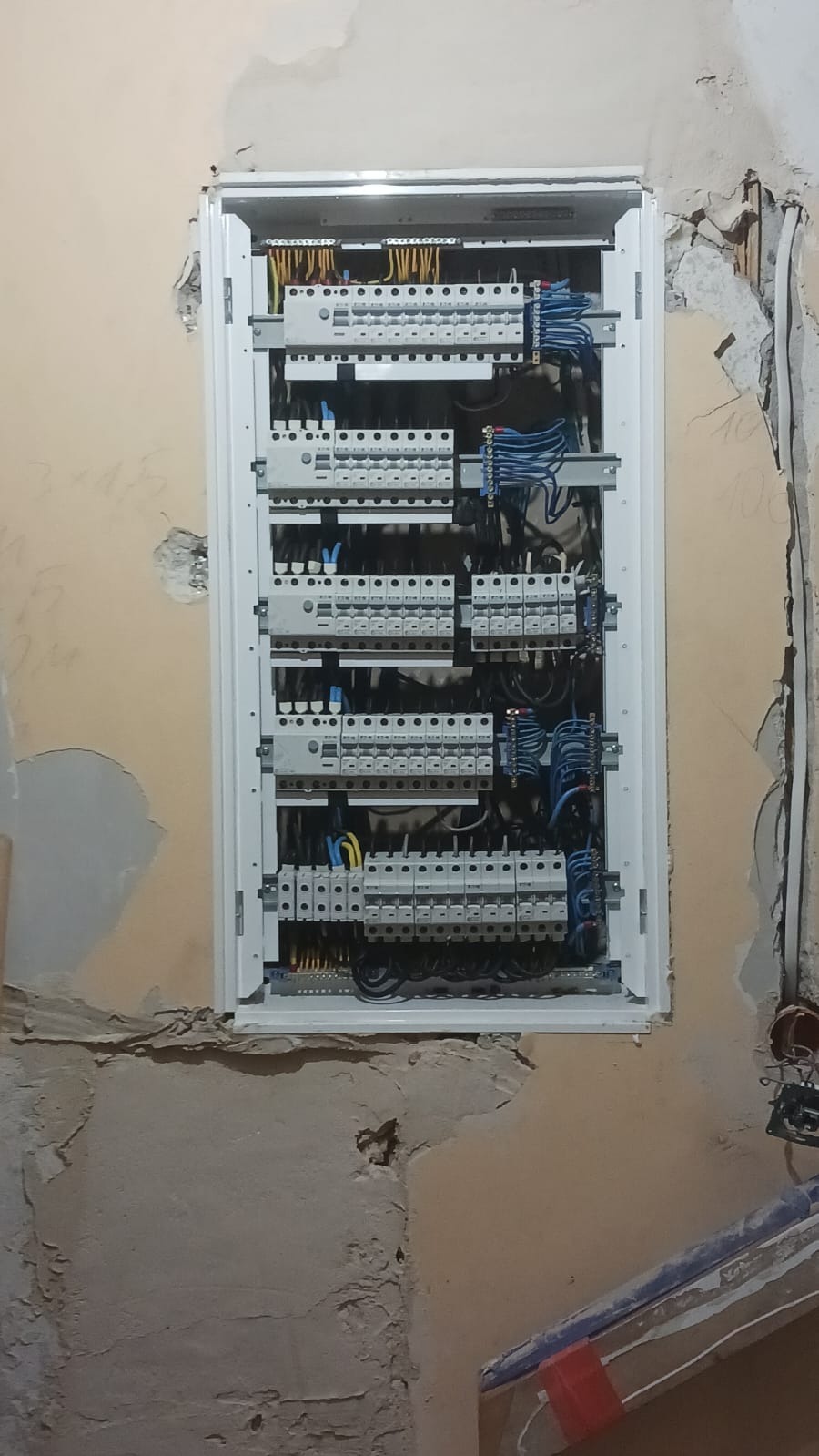 Rozdzielnica elektryczna z licznymi zabezpieczeniami zamontowana w ścianie podczas remontu, widoczne przewody i fragmenty instalacji elektrycznej w surowym stanie.