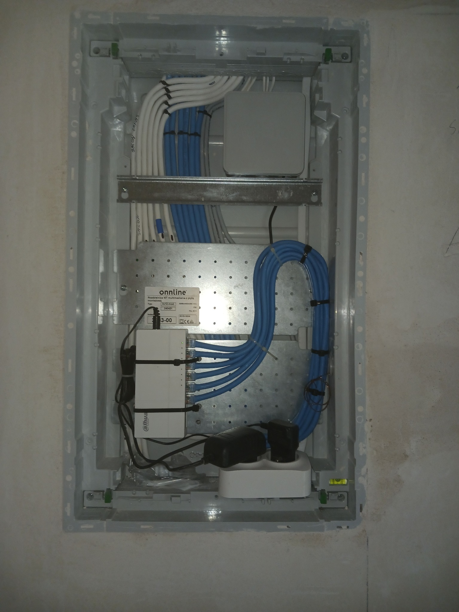 Wewnętrzna skrzynka rozdzielcza z przewodami w kolorach białym i niebieskim, zamontowana w ścianie, widoczna rozdzielnica multimedialna z płytą montażową marki Onnline oraz urządzenia Dahua...