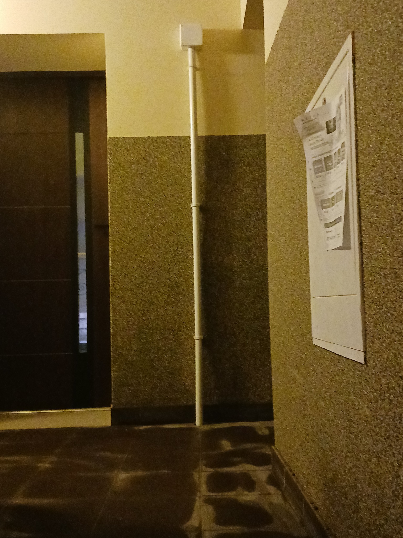 Korytarz z rurą instalacji elektrycznej biegnącą wzdłuż ściany, na której wisi tablica ogłoszeń.