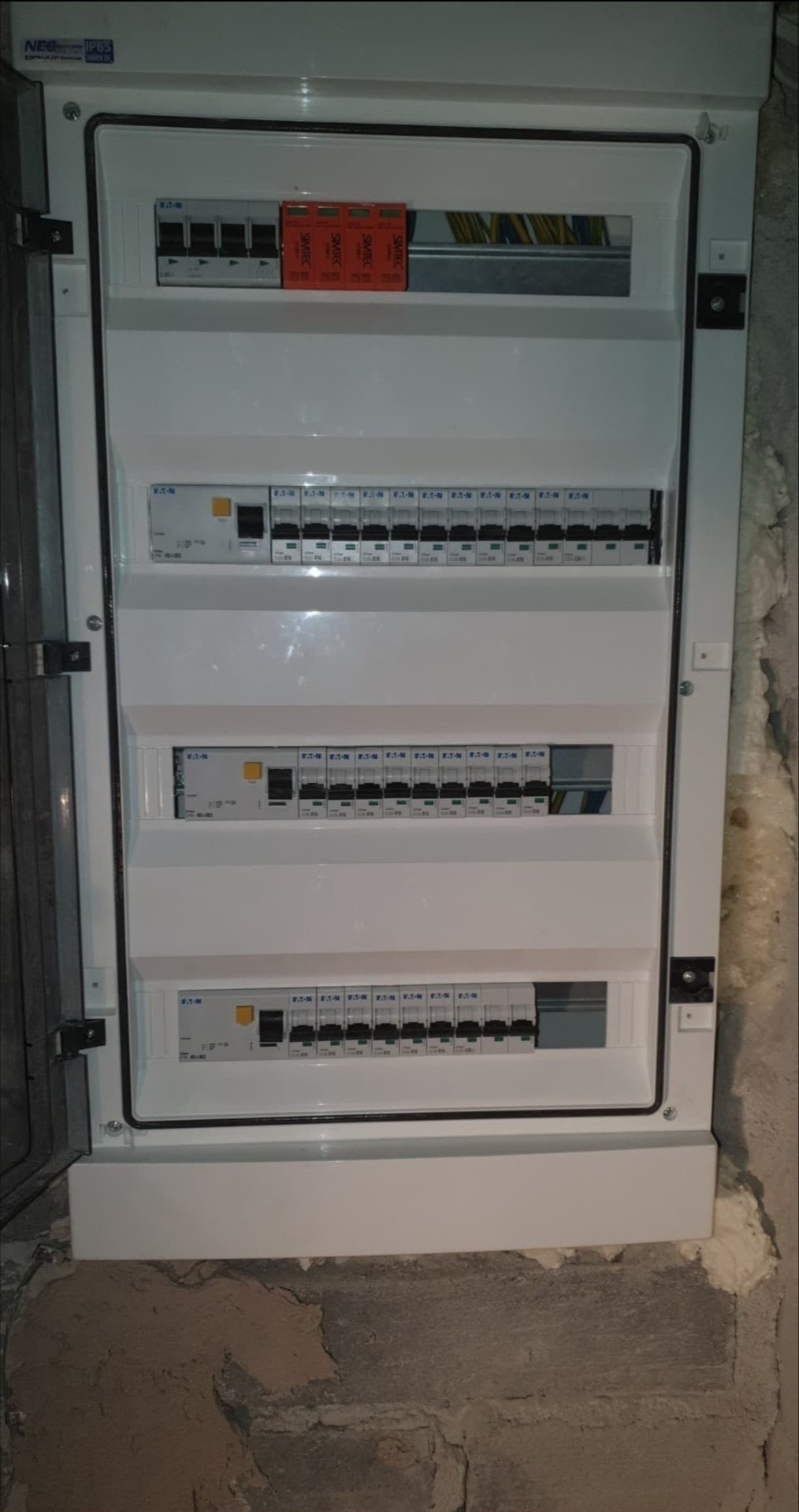 Otwarta skrzynka elektryczna Eaton z widocznymi bezpiecznikami, modułami SIMTEC i przewodami, zamontowana na ścianie z widocznymi śladami pianki montażowej.