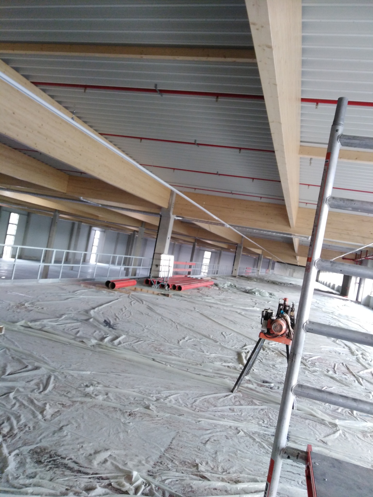 Wnętrze hali produkcyjnej w trakcie budowy z widocznymi elementami konstrukcyjnymi dachu, instalacjami pod stropem, rozłożoną folią ochronną na podłodze, oraz rozstawioną drabiną i agregatem...