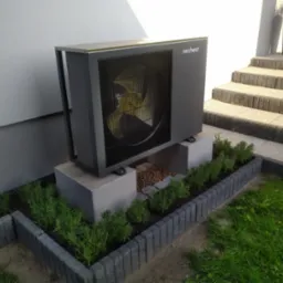 Szara pompa ciepła marki Nesheat, umieszczona na betonowych blokach w ozdobnym ogrodzie z kostki brukowej i niskich roślin, z widocznymi schodami wejściowymi do budynku w tle.