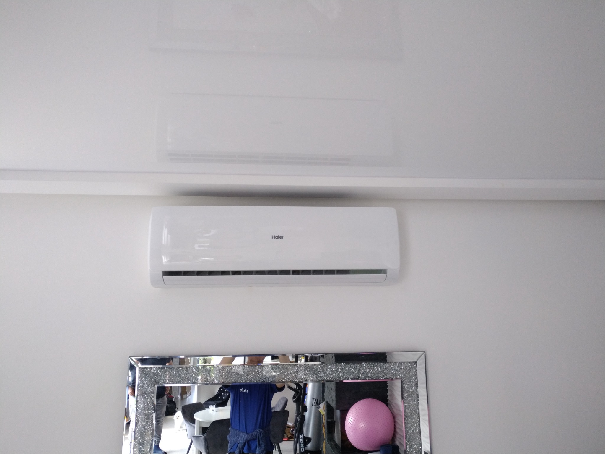 Biały klimatyzator Haier zamontowany na ścianie nad ozdobnym lustrem z kryształkami, odbijającym fragment pomieszczenia z krzesłami i piłką do ćwiczeń.
