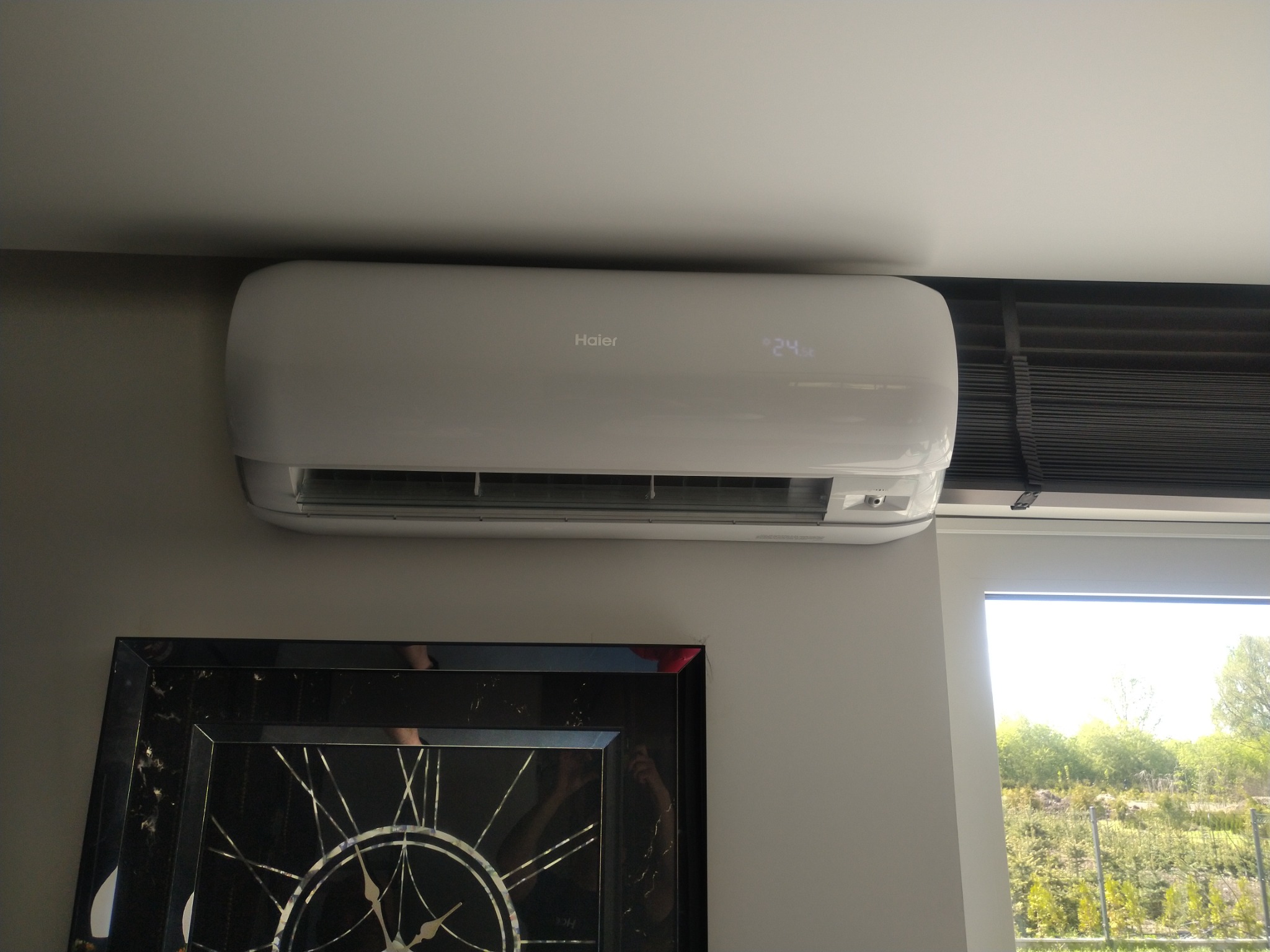 Biały klimatyzator Haier zamontowany pod sufitem, z wyświetlaną temperaturą 24 stopnie, widoczny nad ozdobnym zegarem w czarnej ramie, okno z widokiem na zieleń w tle.