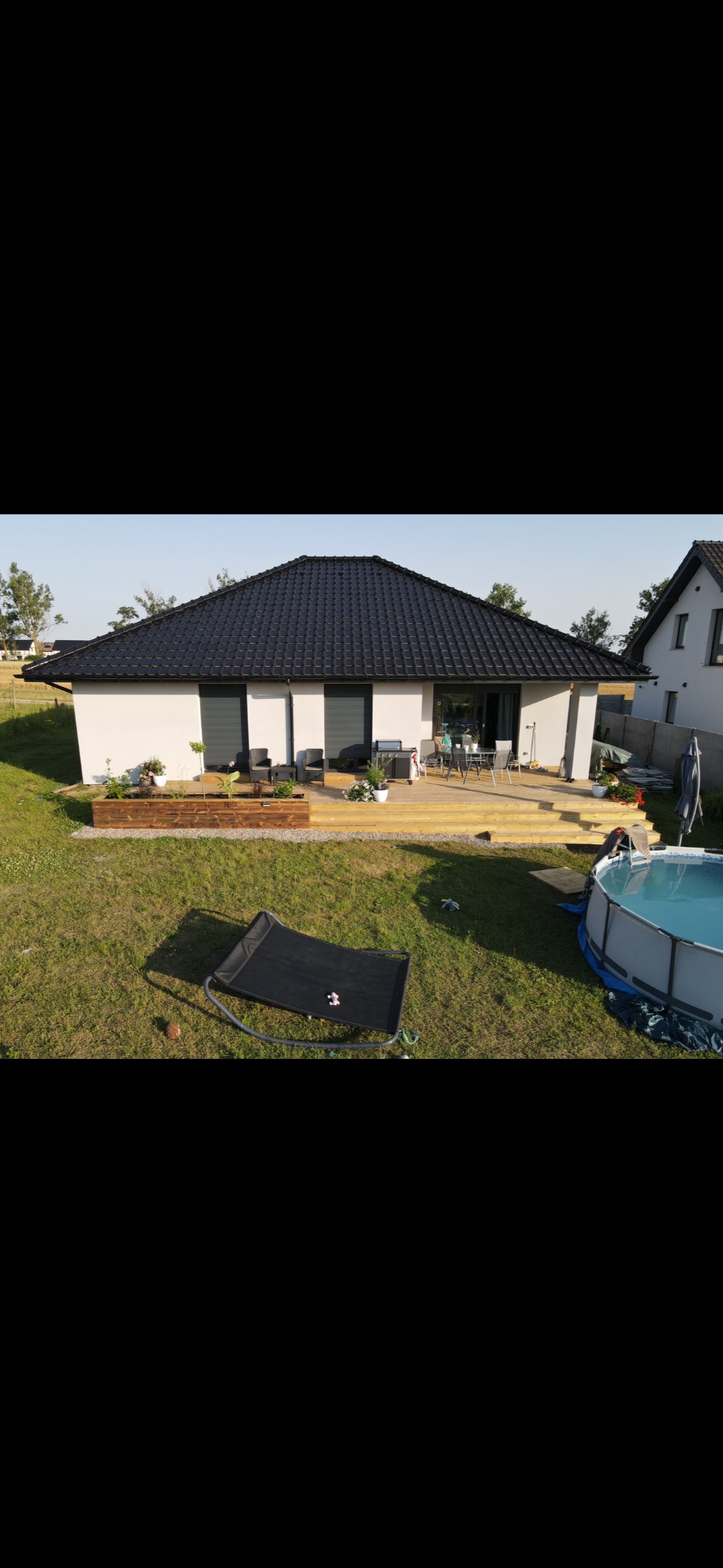 Widok z góry na parterowy dom z czarnym dachem i tarasem, na którym znajdują się meble ogrodowe. Na trawniku basen i leżak.