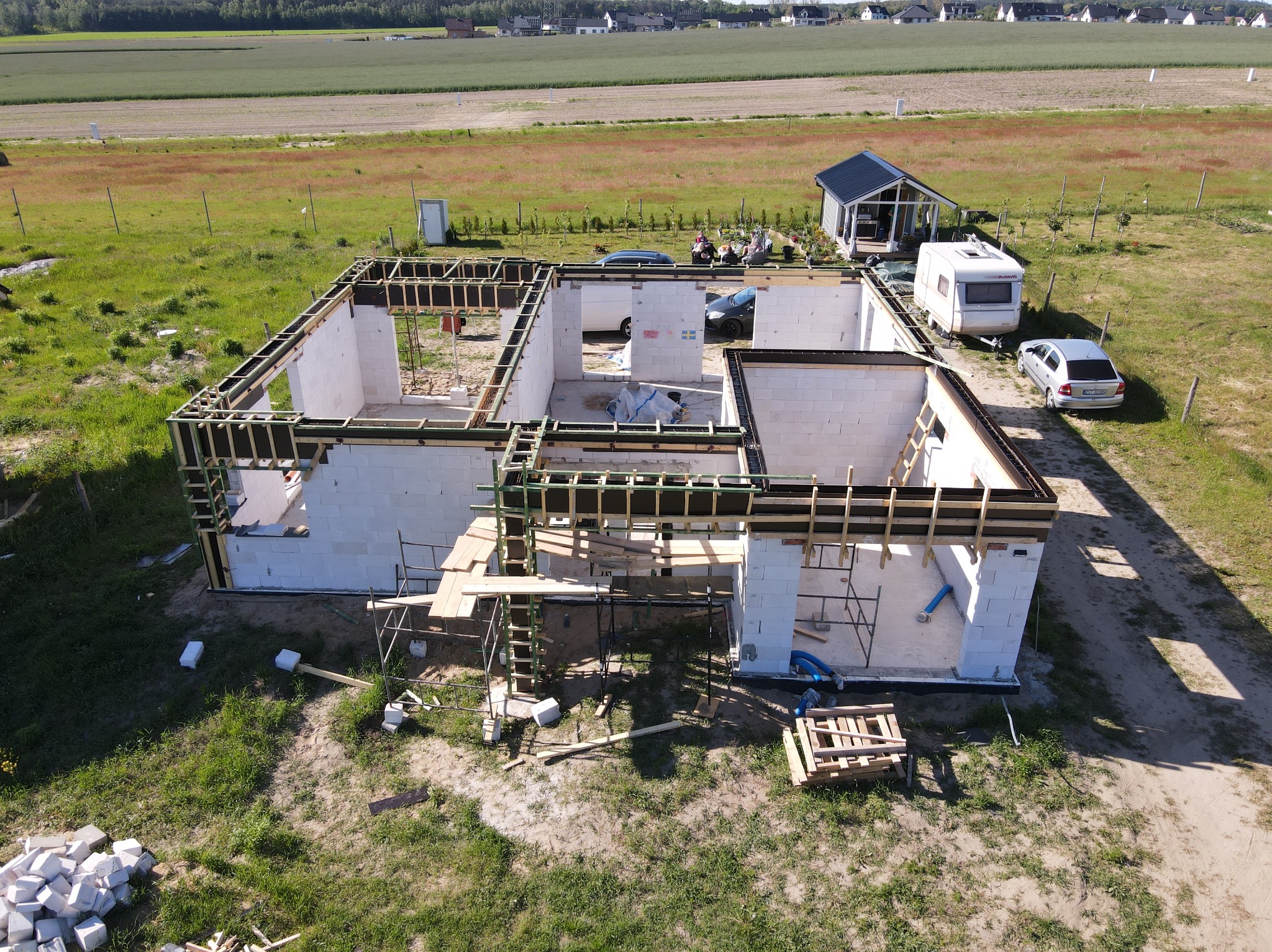 Stan surowy otwarty domu z białych bloczków, widok z drona. Drewniane szalunki na wieńcu. Budowa w Stargardzie, w tle pola i zabudowa jednorodzinna.