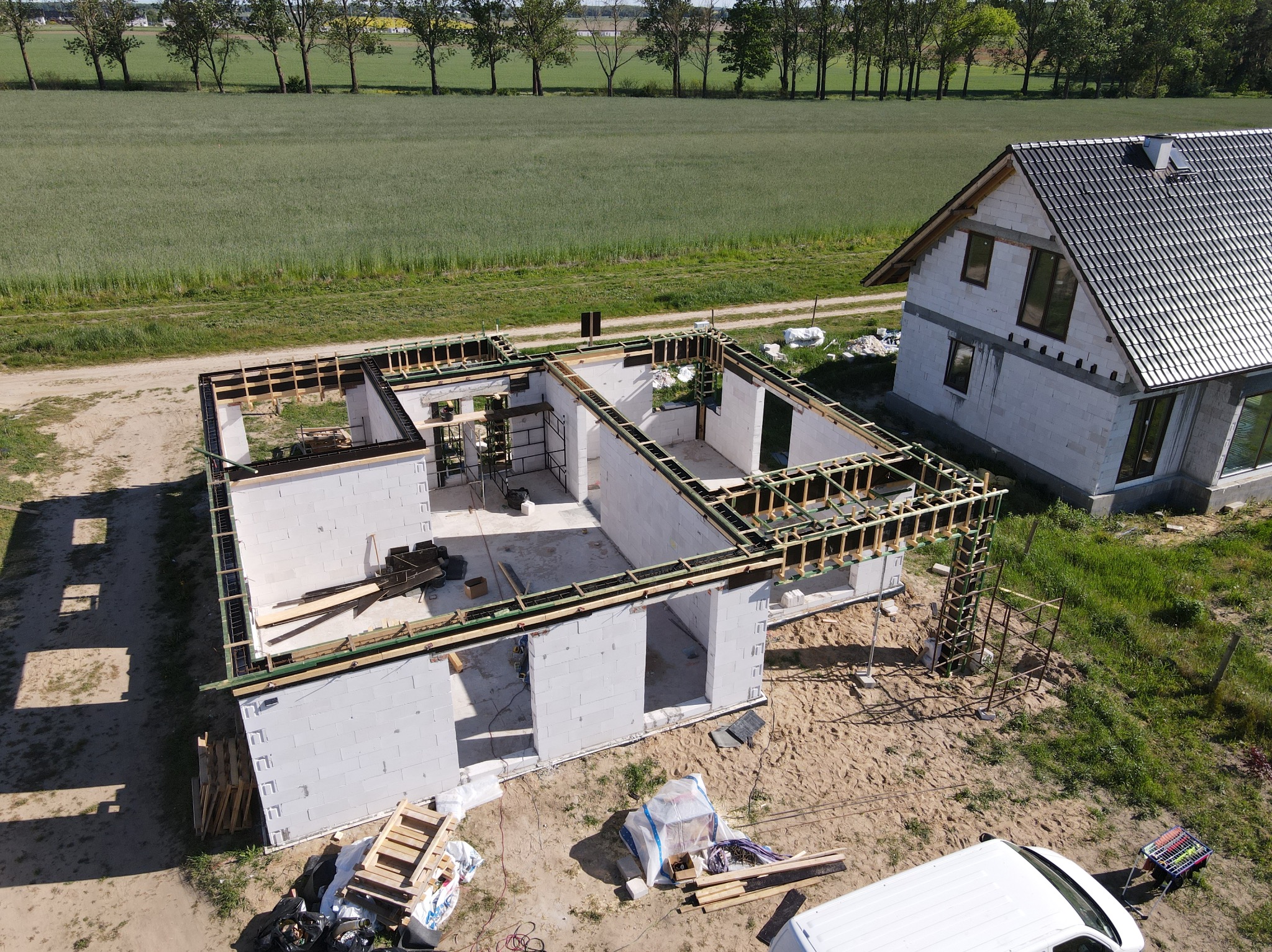 Budowa domu z białych bloczków, widok z góry. Częściowo zadaszony budynek obok, zielone pola w tle. Widoczne drewniane szalunki i elementy konstrukcyjne.