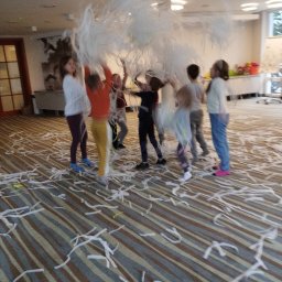 Tygrysek Artur Mokański firma animacyjna - Grupa dzieci rzuca w górę konfetti z białej bibuły w przestronnym pomieszczeniu z wzorzystą wykładziną.