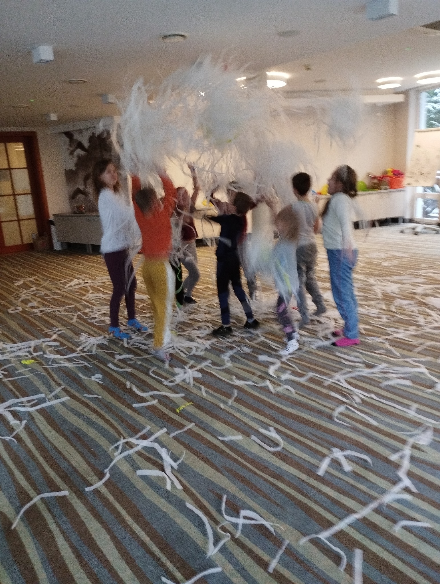 Grupa dzieci rzuca w górę konfetti z białej bibuły w przestronnym pomieszczeniu z wzorzystą wykładziną.