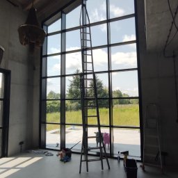 NanoCleaning Polska - Wnętrze z wysokimi oknami po myciu, drabina, narzędzia, widok na zewnątrz. Dwa ozdobne byki przed oknem. Duże okna z czarnymi ramami.