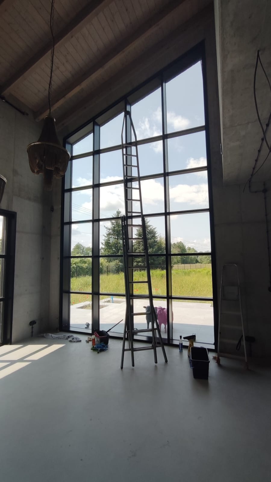 Wnętrze z wysokimi oknami po myciu, drabina, narzędzia, widok na zewnątrz. Dwa ozdobne byki przed oknem. Duże okna z czarnymi ramami.