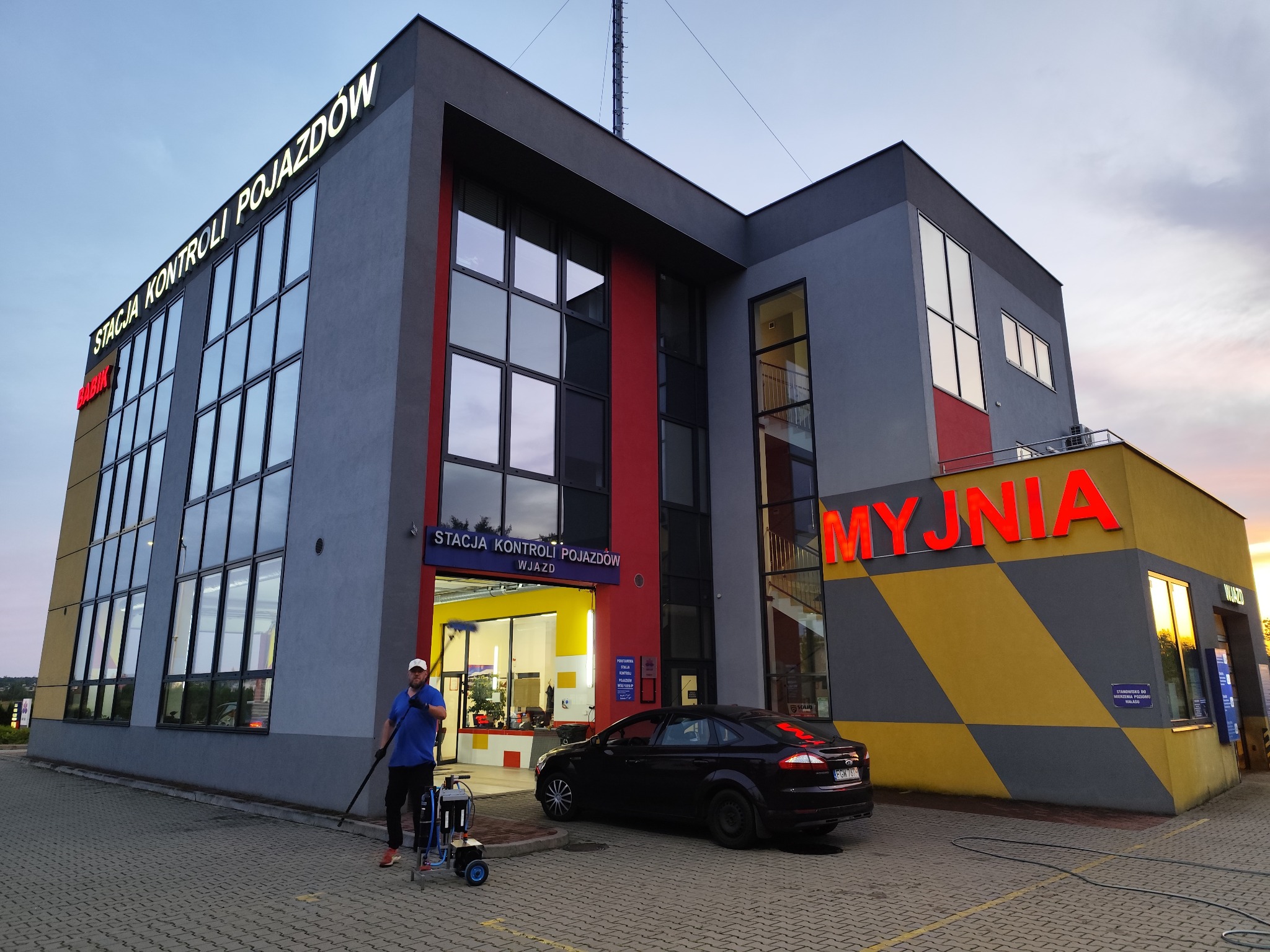 Pracownik myjni samochodowej z urządzeniem do mycia okien na kiju, przed budynkiem stacji kontroli pojazdów z napisem 'MYJNIA' i zaparkowanym autem.
