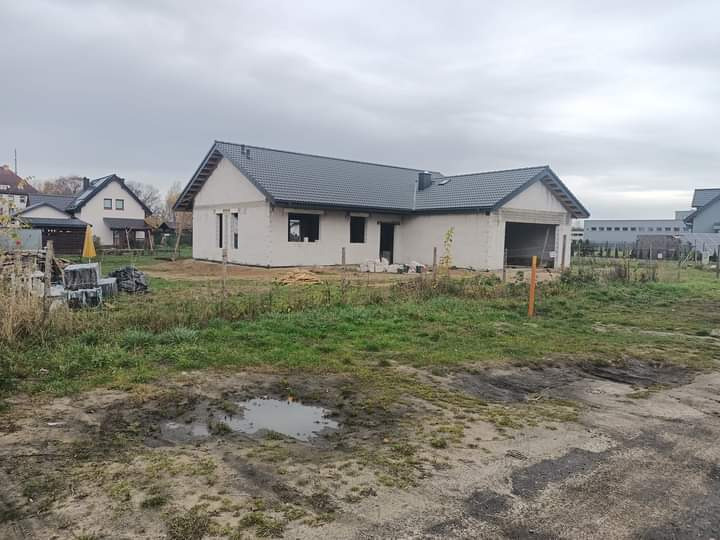 Niedokończony dom jednorodzinny z szarą dachówką, widoczne pustaki i częściowo wykonana elewacja, teren wokół domu niezagospodarowany, pochmurne niebo.