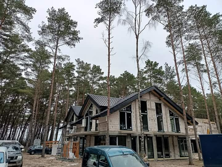 Dom w trakcie budowy z czarną dachówką, otoczony sosnowym lasem, widoczny z poziomu ziemi, z samochodami w tle.