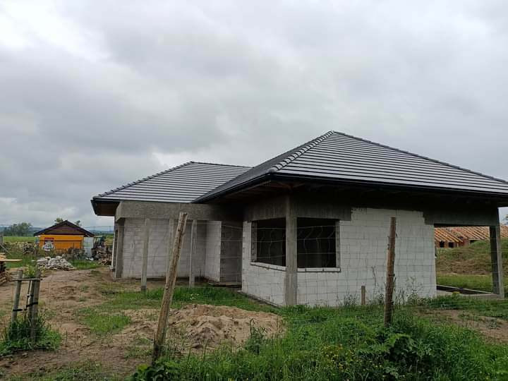 Szary dach z dachówki ceramicznej na surowym budynku w trakcie budowy, widoczne niezadaszone okna i otoczenie placu budowy.