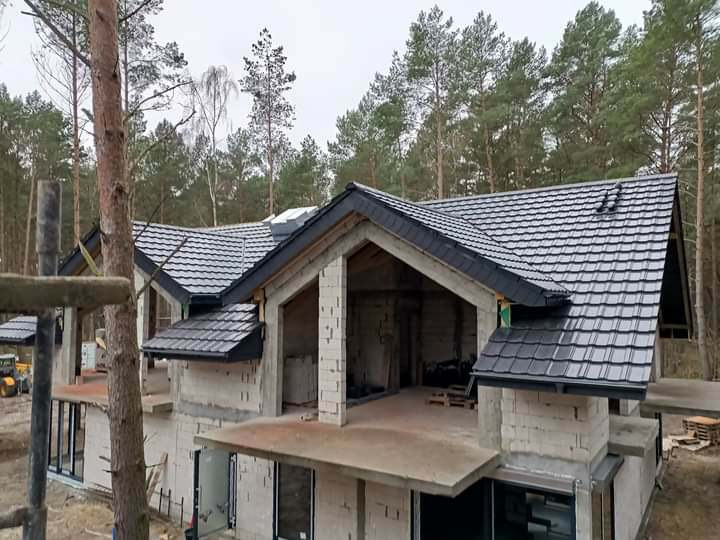 Czarny dach z dachówki ceramicznej na surowym domu w lesie, widoczne fragmenty rusztowania i maszyna budowlana w tle.