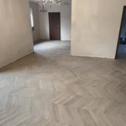 Jasna, drewniana podłoga ułożona w jodełkę w przestronnym, remontowanym wnętrzu. Ściany przygotowane do malowania, widoczny fragment schod&oacute;w i worki z zaprawą w przejściu.