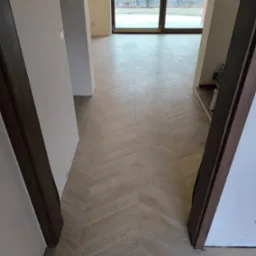 Ułożona w jodełkę podłoga z płytek imitujących drewno, widok z perspektywy przejścia między pomieszczeniami, ściany w stanie surowym, duże okno przesuwne w tle.