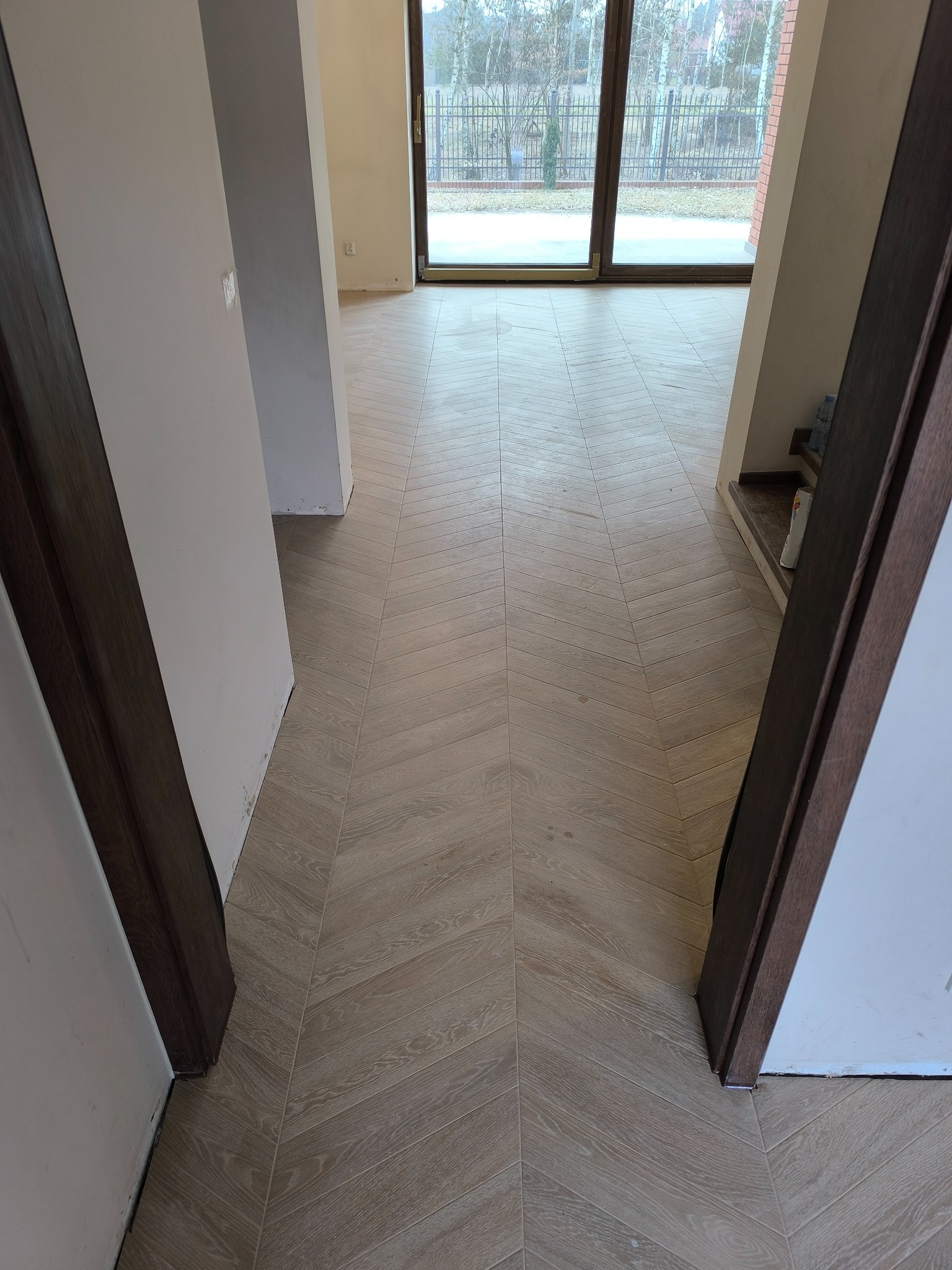 Ułożona w jodełkę podłoga z płytek imitujących drewno, widok z perspektywy przejścia między pomieszczeniami, ściany w stanie surowym, duże okno przesuwne w tle.