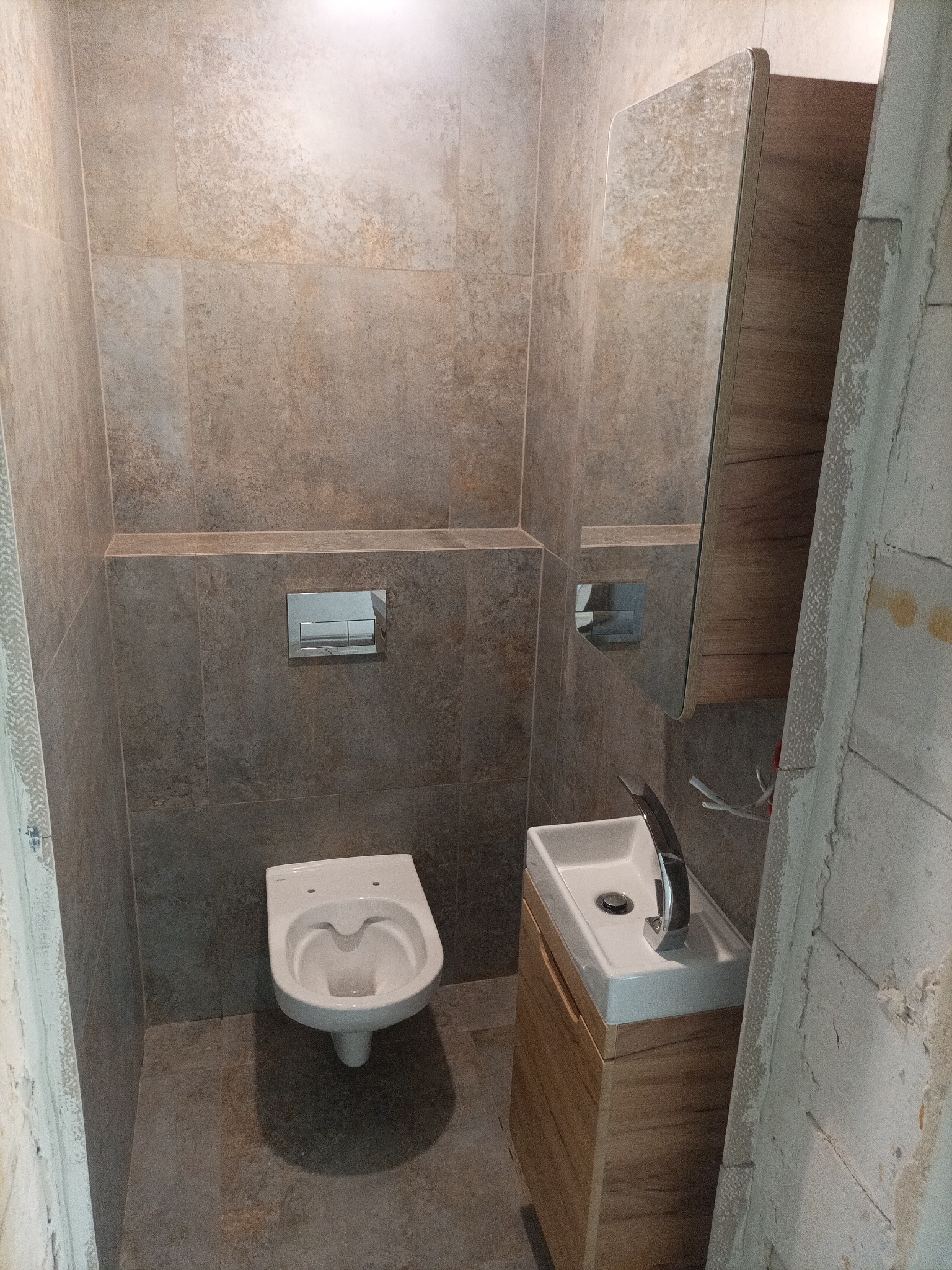 Wnętrze małej łazienki w trakcie remontu z płytkami w odcieniach beżu i brązu, wisząca toaleta, umywalka z szafką z drewnopodobnym wykończeniem i lustro z szafką, widoczne niedokończone ściany.