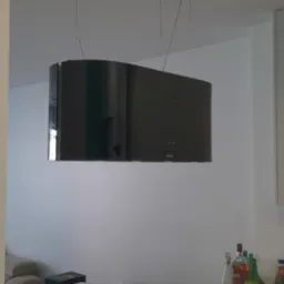 Czarny, designerski okap kuchenny marki MAAN zawieszony na cienkich linkach z czarnymi, prostokątnymi mocowaniami sufitowymi, widoczny nad płytą indukcyjną na drewnianym blacie w jasnej kuchni.