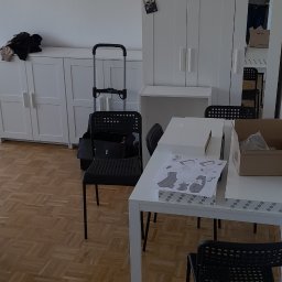 Dariusz Wiśniewski - Rozłożona instrukcja montażu krzesła na białym stole, obok kartonowe pudełko z elementami, w tle białe meble i cztery krzesła, widok z góry.