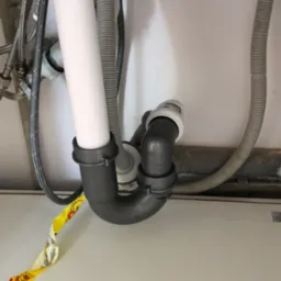 Szare plastikowe rury odpływowe umywalki z syfonem, widoczne podłączenia i elastyczne węże w szafce pod zlewem, fragment żółtej taśmy.