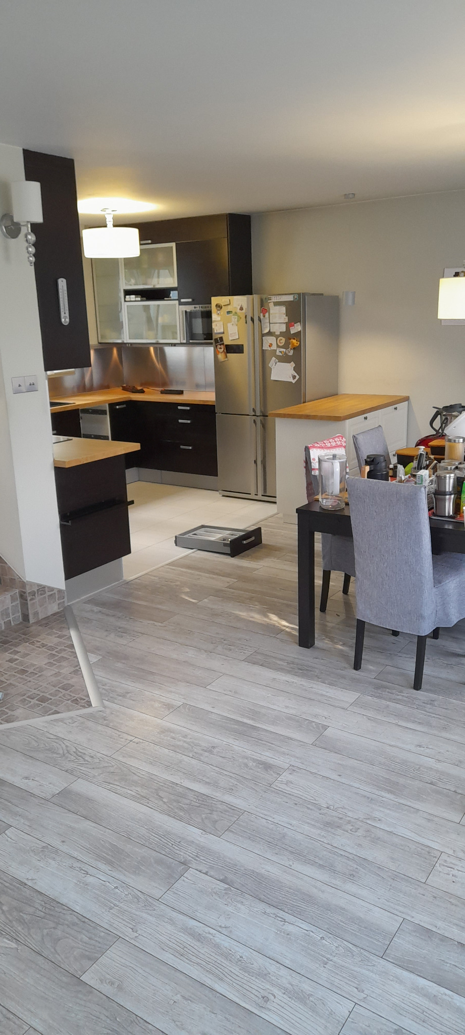 Wnętrze kuchni z elementami montażu: wysunięta szuflada na podłodze, widoczny stół z krzesłami, zabudowa kuchenna z metalowymi elementami i drewnianymi blatami.