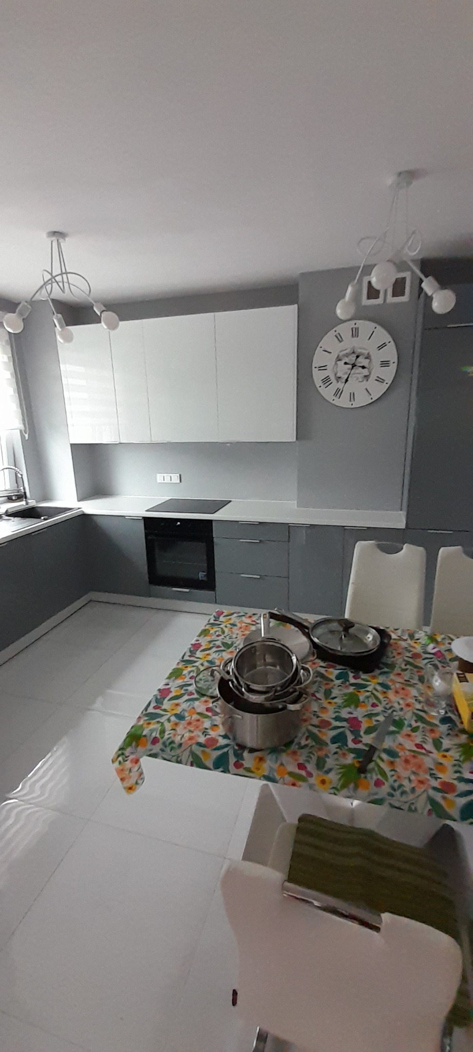 Szaro-biała kuchnia z nowoczesnym oświetleniem i dużym zegarem ściennym, stół nakryty kwiecistym obrusem z garnkami i patelniami.